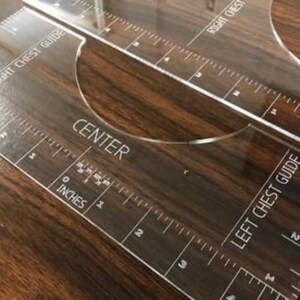 May include: Clear acrylic chest guide with inch measurements and the words "RIGHT CHEST GUIDE", "CENTER", and "LEFT CHEST GUIDE" printed in white. The guide has a curved cutout and is placed on a dark wood surface.