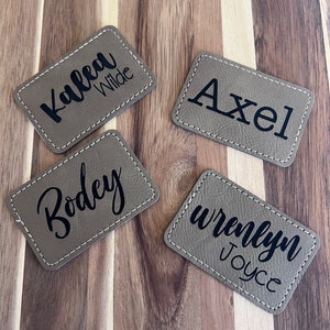 May include: Four rectangular, tan-colored name tags with white stitching around the edges. Each tag has a different name in black script: Kalea Wilde, Axel, Bodey, and Wrenlyn Joyce. The tags are arranged on a wooden surface.
