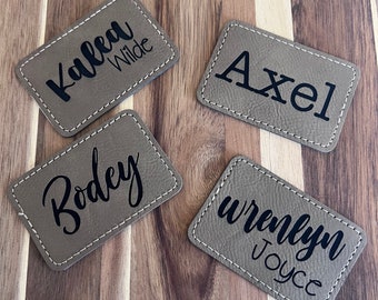 May include: Four rectangular, tan-colored name tags with white stitching around the edges. Each tag has a different name in black script: Kalea Wilde, Axel, Bodey, and Wrenlyn Joyce. The tags are arranged on a wooden surface.