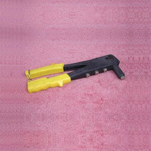 May include: A black and yellow rivet gun tool. The tool has yellow handles and a black body with silver rivets. The tool is designed for fastening materials together using rivets. The tool is on a pink background.