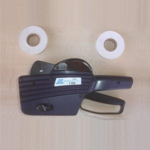 May include: A dark blue price labeler with a black handle and a white label that reads "IMS 18B". Two rolls of white label tape are positioned above the labeler.