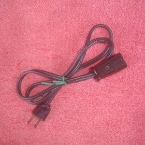 May include: A black electrical power cord with a two-prong plug and a female connector. The cord is looped and secured with a green rubber band. The background is a textured, red surface.