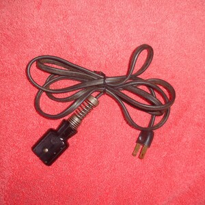 May include: A black electrical cord with a two-prong plug and a female connector with a spring. The cord is coiled and tied, set against a red background.