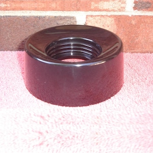 May include: A glossy black object with a threaded interior. The object is cylindrical with a wide, flat top and a rounded base. The background features a brick wall and a pink surface.
