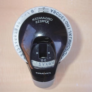 May include: A black and gray label maker with the text "ORGANISER XPRESS" and "DYMO". The label maker has a dial with letters and numbers, and a display showing "SYXWV".