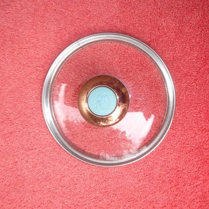 May include: A clear glass lid with a silver rim and a copper-colored center knob. The knob has a light blue insert with the letters "PD" in white. The lid is on a red background.