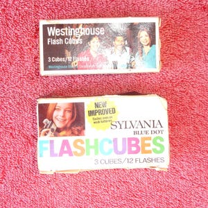 May include: Two boxes of flash cubes. The top box is white with blue and red text that reads "Westinghouse Flash Cubes 3 Cubes/12 Flashes". The bottom box is white with blue, red, green, and yellow text that reads "SYLVANIA BLUE DOT FLASHCUBES 3 CUBES/12 FLASHES". The bottom box also has a yellow rectangle with black text that reads "NEW IMPROVED flashes even on weak batteries".