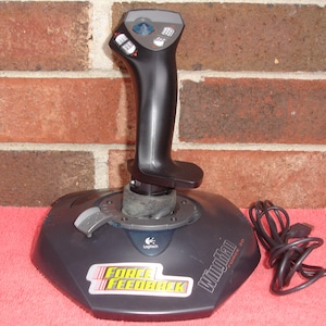 May include: A black Logitech WingMan Force 3D joystick with a black cable. The joystick has a black handle and a base with the words "FORCE FEEDBACK" and "WingMan FORCE 3D". The background is a red surface and a brick wall.
