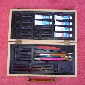 Puede incluir: Un estuche de pintura de artista de madera abierto, que contiene tubos de pintura y pinceles. Los tubos de pintura son blancos con etiquetas azules y rosas. Los pinceles tienen mangos de madera y cerdas de varios colores. El set está diseñado para artistas.