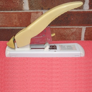 May include: A vintage stapler with a cream-colored handle and a white base. The stapler has a curved handle and a red metal component. The background features a brick wall and a pink surface.