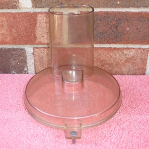 May include: A clear, light-brown food processor part with a circular base and a tall, cylindrical top. The base has a small, rectangular protrusion and a hinged latch. The part is made of transparent plastic and sits on a pink towel.
