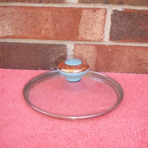 May include: A clear glass lid with a silver rim and a decorative knob. The knob has a light blue base with a copper-colored top. The lid is resting on a pink surface, with a brick wall in the background.