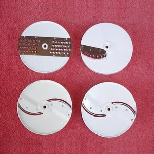 May include: Four white, circular food processor blades with stainless steel cutting surfaces. Two blades have grating surfaces, and two have slicing blades. The blades are arranged on a red background.
