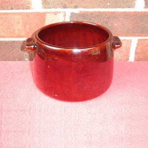 May include: A glossy, reddish-brown ceramic pot with two small handles. The pot has a wide opening and a rounded body. The background features a brick wall and a pink surface.