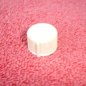 May include: A white plastic bottle cap with a ribbed side and a flat top. The cap is cylindrical and sits on a textured, pink surface. The cap is a common item used for sealing bottles.