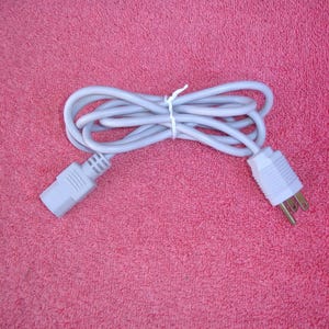 May include: A coiled, light gray power cord with a standard three-prong plug on one end and a rectangular connector on the other. The cord is secured with a white cable tie, set against a pink background.