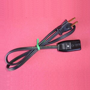 Peut inclure: Un cordon d'extension électrique noir avec une prise à deux broches et une prise femelle. Le cordon est enroulé et fixé avec une attache torsadée verte. Le texte "18 AWG 2/C TYPE HPN HEATER" est imprimé sur le cordon.
