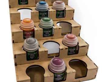 May include: A tiered, brown cardboard paint organizer holding several Citadel Shade paint pots. The pots are arranged in a stepped fashion, with various colors visible, including sepia, drakenhof nightshade, and agrax earthshade.