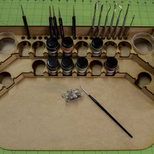 May include: A wooden craft organizer with various tools and supplies. It features paint bottles, brushes, and sculpting tools. The organizer has a large flat surface for projects and two small containers.