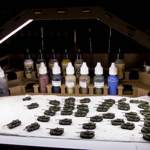 Pode incluir: Uma bancada de trabalho com uma variedade de frascos de tinta e tanques em miniatura. A bancada de trabalho tem uma fonte de luz acima, iluminando os tanques.