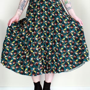 May include: A pleated midi skirt with a teal background and a repeating floral pattern in yellow, brown, and black. The skirt is paired with a black top and black ankle boots. The model is holding the skirt out.