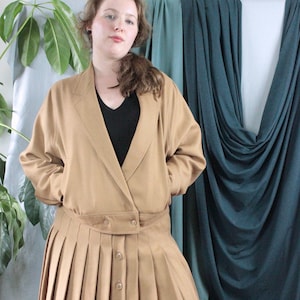 Peut inclure: Une jupe plissée beige avec une veste assortie. La veste a une fermeture à double boutonnage et un col. La jupe a une fermeture à boutons et est plissée.