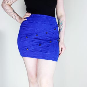 May include: A vibrant blue mini skirt with a textured fabric and a wrap-around design. The skirt is adorned with small, gold-colored buttons. The garment is a stylish piece of clothing.