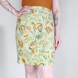 Op de afbeelding: Lichtgroene potloodrok met een bloemenpatroon in bruin-, oranje- en groentinten. De rok komt tot op de knie en heeft een getailleerd silhouet. Het bloemmotief is gedetailleerd en bedekt de hele rok.