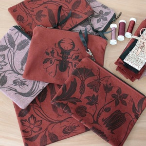 Puede incluir: Cinco bolsas de tela roja con diseños impresos en negro. Los diseños incluyen un escarabajo ciervo volante, una flor y una hoja. Las bolsas están hechas de una tela suave y texturizada.
