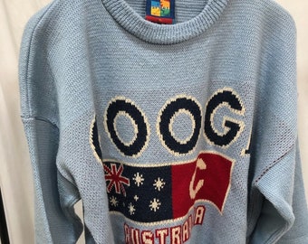 May include: A light blue knitted sweater with a large "COOGI" logo in white and navy blue lettering. The sweater features a red, white, and blue striped band at the bottom and a red, white, and blue Australian flag design with stars and a kangaroo.