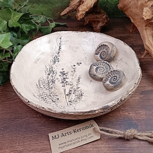 Könnte beinhalten: Eine handgefertigte Keramikschale mit cremefarbener Glasur und floralem Design. Die Schale enthält drei kleine, runde, fossilartige Steine. Ein Etikett mit dem Text "MJ-Arts-Keramik" ist angebracht. Die Schale hat einen Durchmesser von ca. 20 cm.