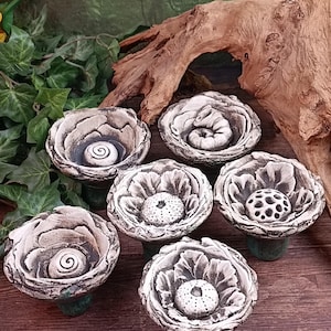 Op de afbeelding: Zes handgemaakte keramische bloemvormige kommen met een getextureerde, gebroken witte afwerking. Elke kom heeft een uniek middenontwerp, sommige met spiraalpatronen en andere met getextureerde bollen. De kommen staan op groene voetstukken.
