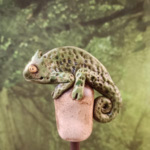 May include: A ceramic chameleon figurine in shades of green and brown, perched atop a small, cylindrical post. The chameleon has detailed spots and a curled tail. The background is a blurred green, suggesting a natural setting.