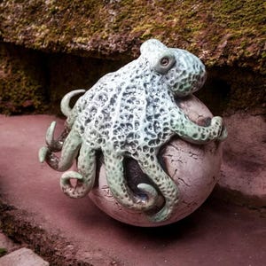 Könnte beinhalten: Eine Keramik-Oktopus-Skulptur in Grün- und Weißtönen, die auf einer strukturierten, kugelförmigen Basis ruht. Der Oktopus hat detaillierte Tentakel und einen strukturierten Körper. Die Skulptur steht vor einem natürlichen Hintergrund.