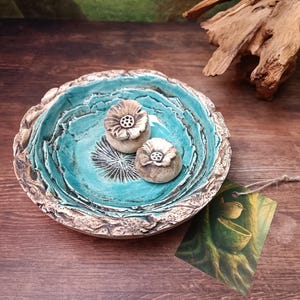 Op de afbeelding: Een ronde keramische kom met een textuur die op schors lijkt aan de buitenkant en een fel turquoise geglazuurde binnenkant. In de kom liggen twee kleine, beige, bloemvormige keramische objecten. Een rechthoekig label is bevestigd met touw.