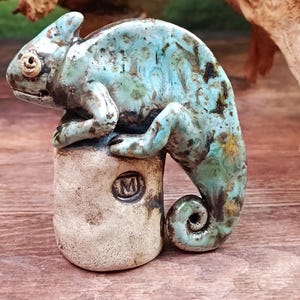 May include: A ceramic chameleon sculpture in shades of turquoise, brown, and gold. The reptile is perched on a cylindrical base with an embossed "M" logo. The sculpture has a glossy finish and is set against a natural wood background.