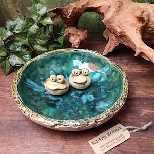 Könnte beinhalten: Eine runde Keramikschale mit glänzender, smaragd- und türkisgrüner Glasur und einem strukturierten, rindenartigen Rand. Zwei kleine, beige, modellierte Froschfiguren mit großen Augen sitzen in der Schale. Ein beiges Etikett trägt MJ-Arts-Keramik.