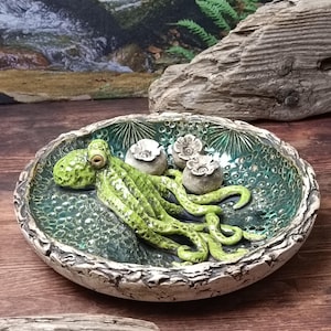 Könnte beinhalten: Eine handgefertigte Keramikschale mit einer grünen Oktopus-Skulptur und drei kleinen blütenartigen Dekorationen. Die Schale hat einen strukturierten, cremefarbenen Rand und einen gesprenkelten grünen Innenraum. Der Oktopus ist leuchtend grün.