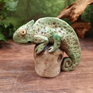 May include: A ceramic chameleon figurine in shades of green and brown, perched atop a textured, rounded base. The reptile has detailed scales and a curled tail. The figurine is a decorative item.