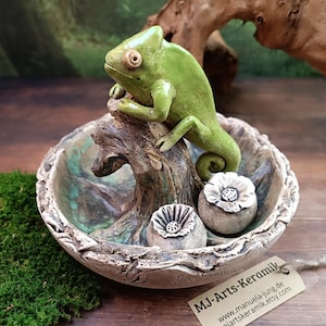 Könnte beinhalten: Handgefertigte Keramikskulptur mit einem grünen Chamäleon, das auf einem Ast in einer dekorativen Schale sitzt. Die Schale hat ein strukturiertes, erdiges Design mit zwei kleinen blütenförmigen Akzenten. Die Skulptur ist vor einem natürlichen Hintergrund platziert.