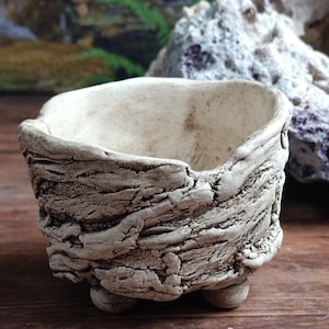 Op de afbeelding: Een kleine, gebroken witte keramische plantenbak met een getextureerde, schorsachtige buitenkant. De plantenbak heeft een afgeronde vorm, ondersteund door drie kleine, afgeronde voetjes. De binnenkant is glad, in tegenstelling tot de ruwe buitenkant.