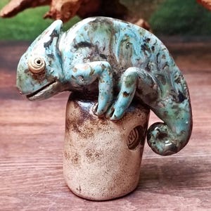 May include: A ceramic chameleon sculpture in shades of blue, green, and brown, perched atop a cylindrical base. The sculpture has detailed features and a textured finish, showcasing the artisan's skill. The base has a textured, earthy appearance.