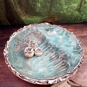 Op de afbeelding: Een handgemaakte keramische kom met een turquoise en zilverkleurige afwerking. De kom heeft een textuur en drie kleine, bloemvormige ornamenten. De rand heeft een getextureerde, zilveren rand. De kom wordt gepresenteerd op een houten oppervlak.