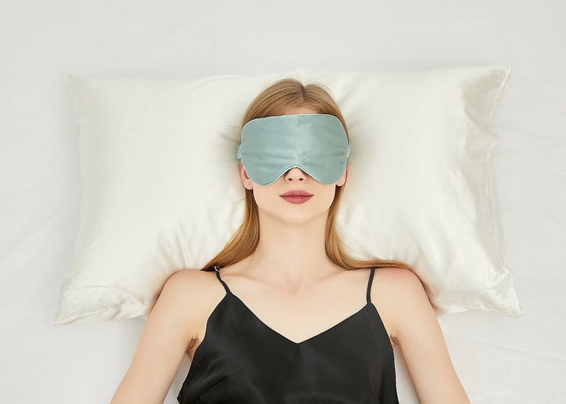 May include: A person wearing a light blue sleep mask lies on a white pillow with a black tank top.