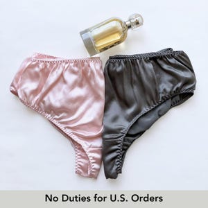 May include: Two pairs of satin knickers in pink and grey, with a perfume bottle in the background. The knickers have a high-waisted design with a ruffled edge. The text at the bottom reads "No Duties for U.S. Orders."