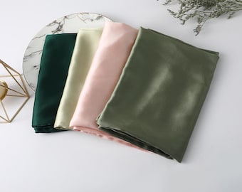 May include: Four folded rectangular fabric pieces with a smooth, satin-like finish. The colors include dark forest green, pale cream, soft pastel pink, and muted olive green. The fabric is arranged in a layered, overlapping stack.