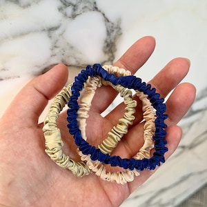 Puede incluir: Tres scrunchies para el pelo en una mano. Uno es azul marino, uno es beige y uno es beige claro.