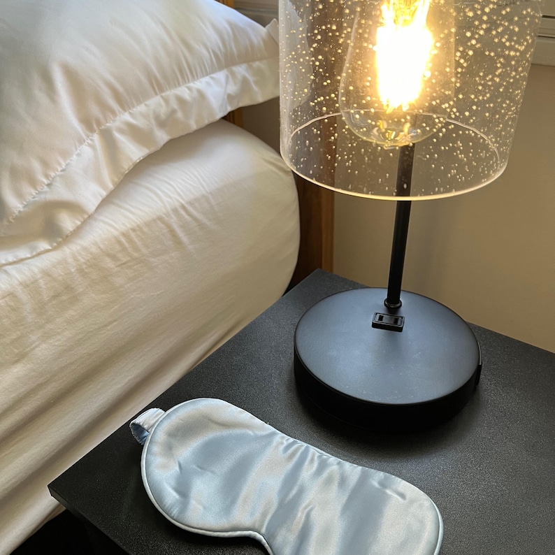 May include: A light blue silk sleep mask on a black nightstand next to a black lamp with a clear glass shade. The lamp is turned on and the light is shining.