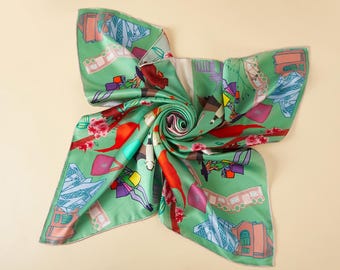 May include: A square silk scarf in a light green hue featuring a colorful print of stylized buildings, pink cherry blossoms, and abstract figures. The design includes geometric architectural shapes, red bird-like forms, and various bags.