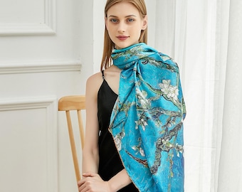 May include: A square silk scarf featuring a vibrant turquoise blue background with a detailed print of white almond blossoms on dark brown branches. The scarf is draped over the shoulder, showcasing a smooth texture and a colorful artistic pattern.
